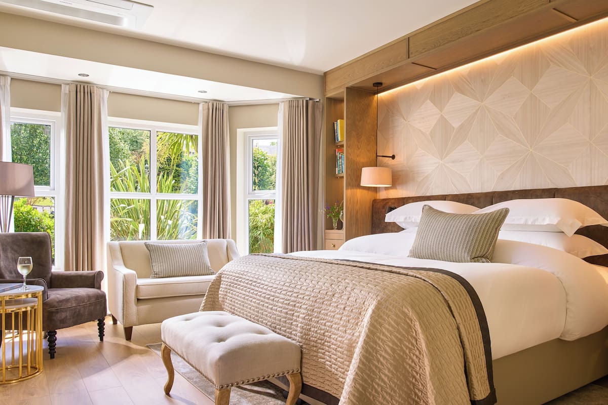 Une chambre d’hôtel dotée d’un grand lit, d’un coin salon avec un fauteuil et un pouf, d’une table de chevet avec une lampe, et offrant une vue sur la verdure par les fenêtres.