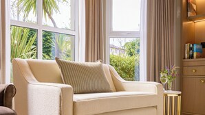 A living room with a beige sofa, a small side table with a vase, and a wooden cabinet with books. - Brook Lane Hotel (Kenmare)