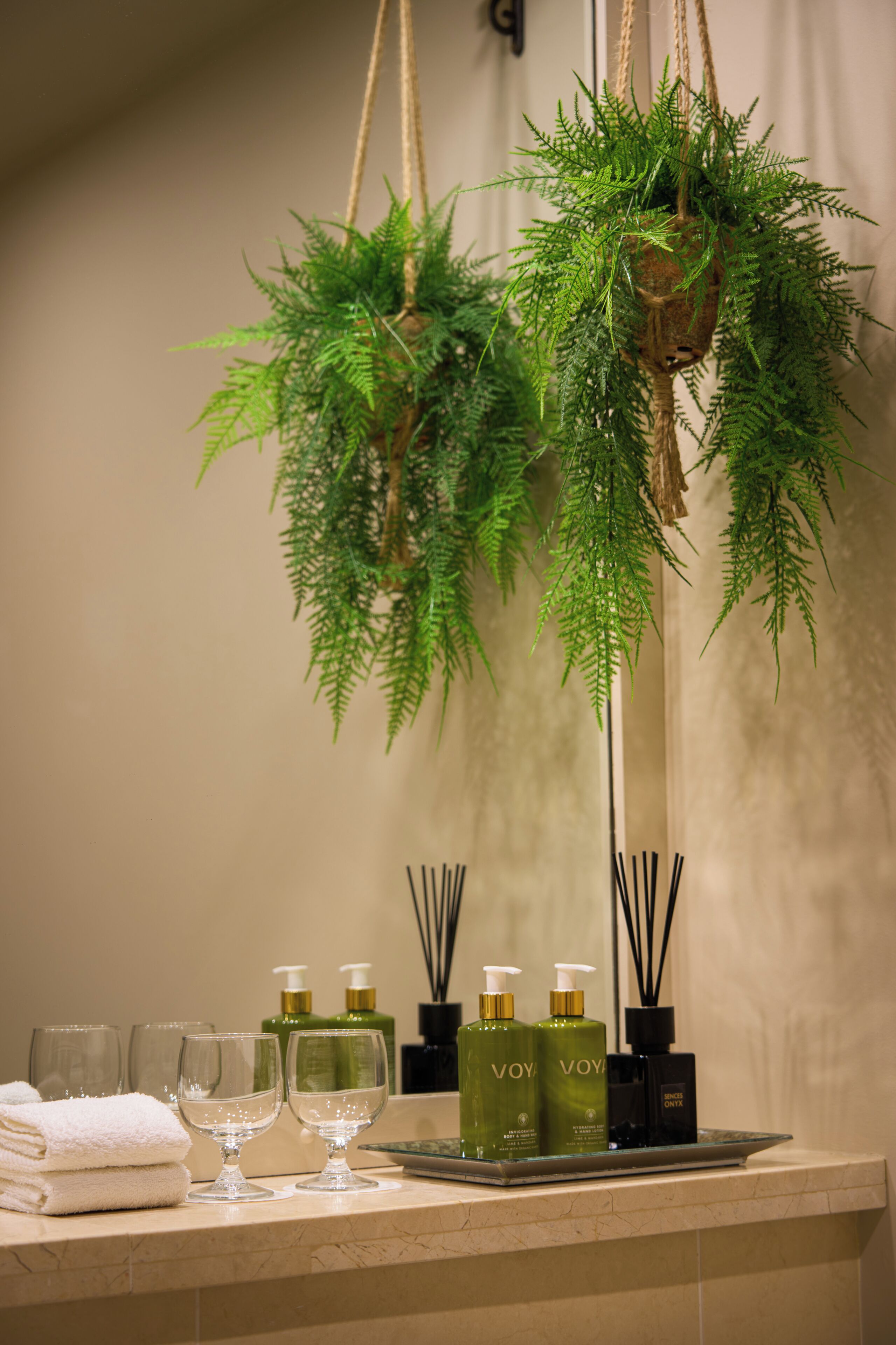 A bathroom with a sink, two green bottles labeled 'VOYA,' a glass, a towel, and a hanging plant.