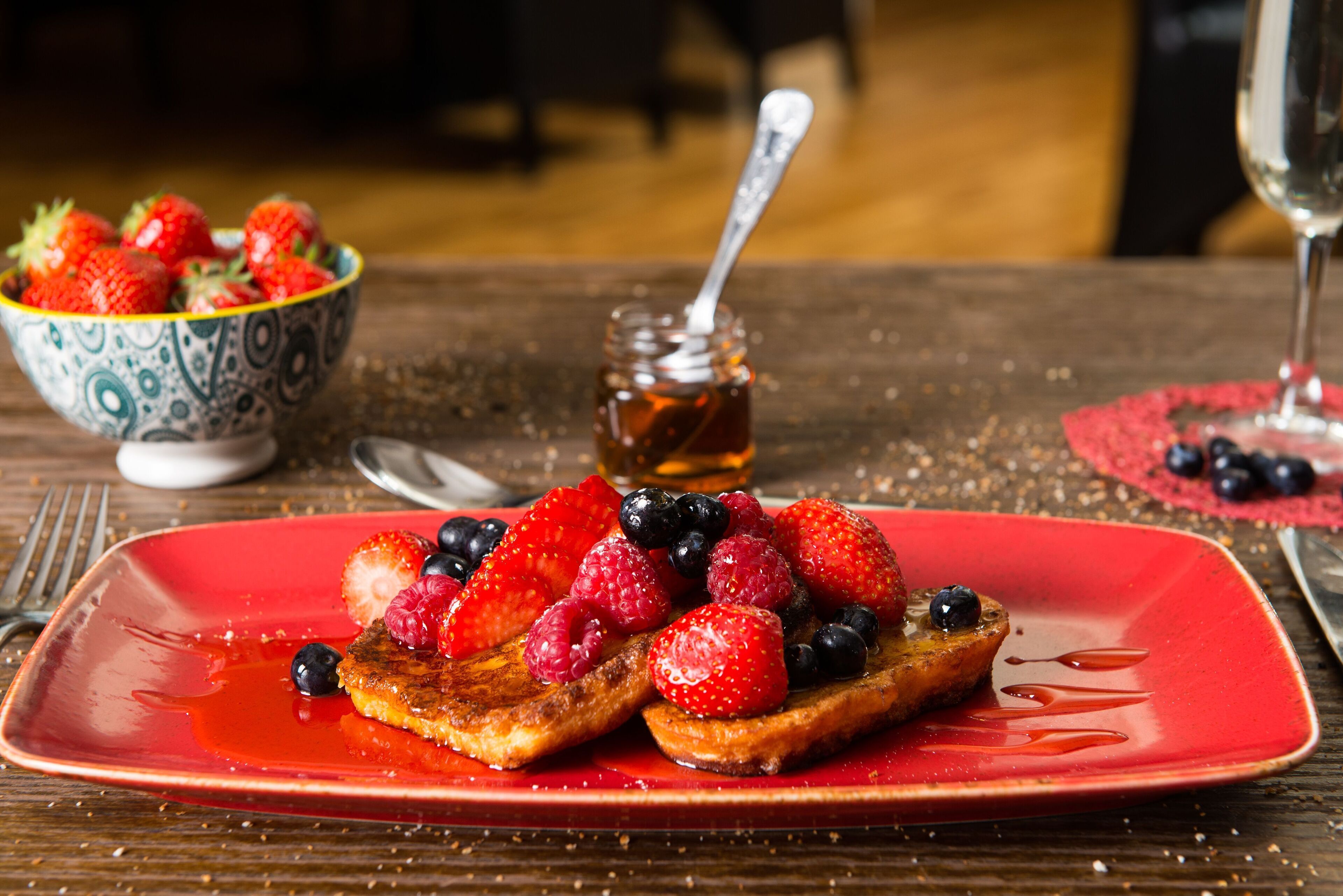 A breakfast plate with French toast, strawberries, and blueberries, a bowl of mixed berries, and a jar of honey on a wooden table.