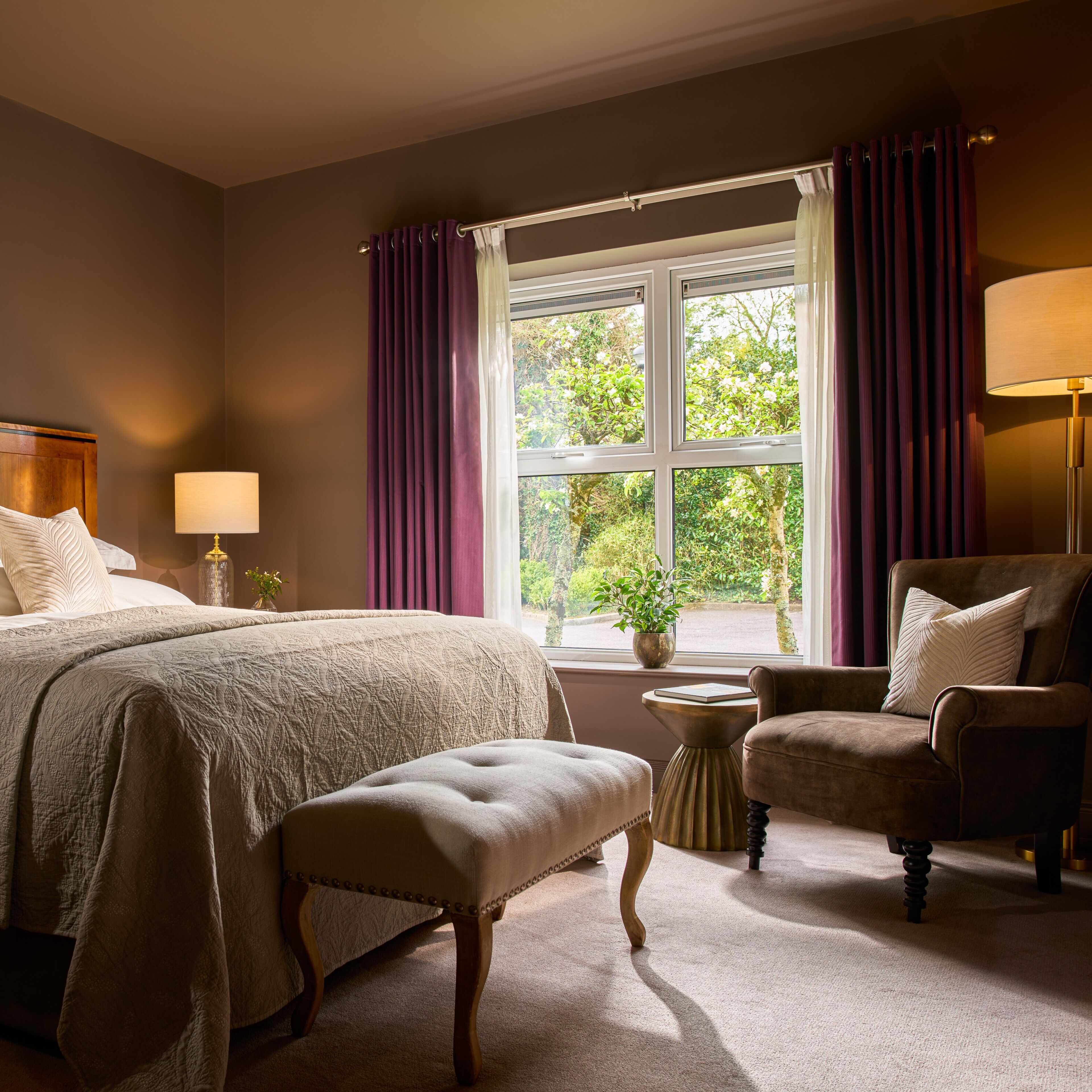 Een slaapkamer met een bed, een stoel, een lamp, een raam met gordijnen en uitzicht op groen buiten.