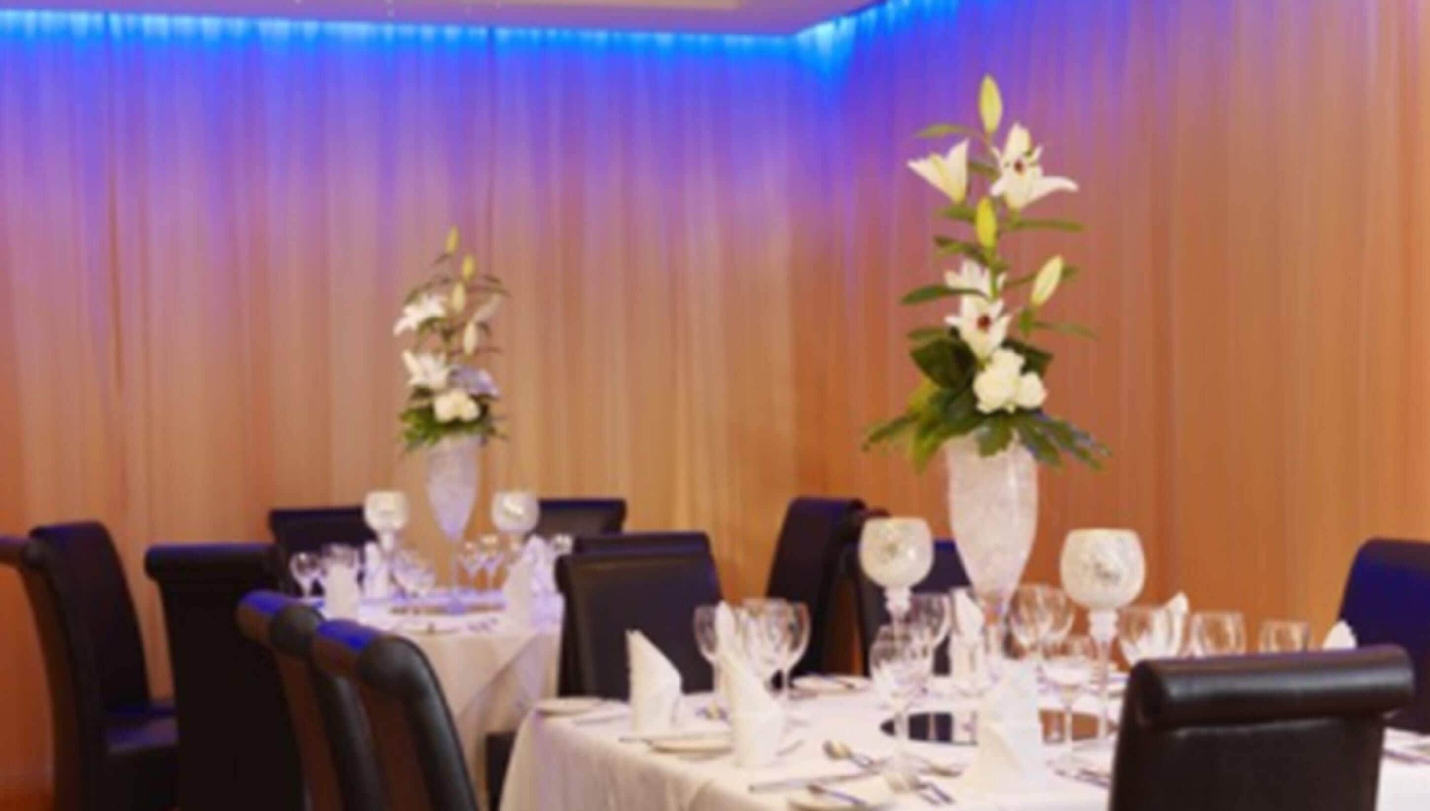 A dining area with tables set for a formal event, featuring tall floral centerpieces and white tablecloths.