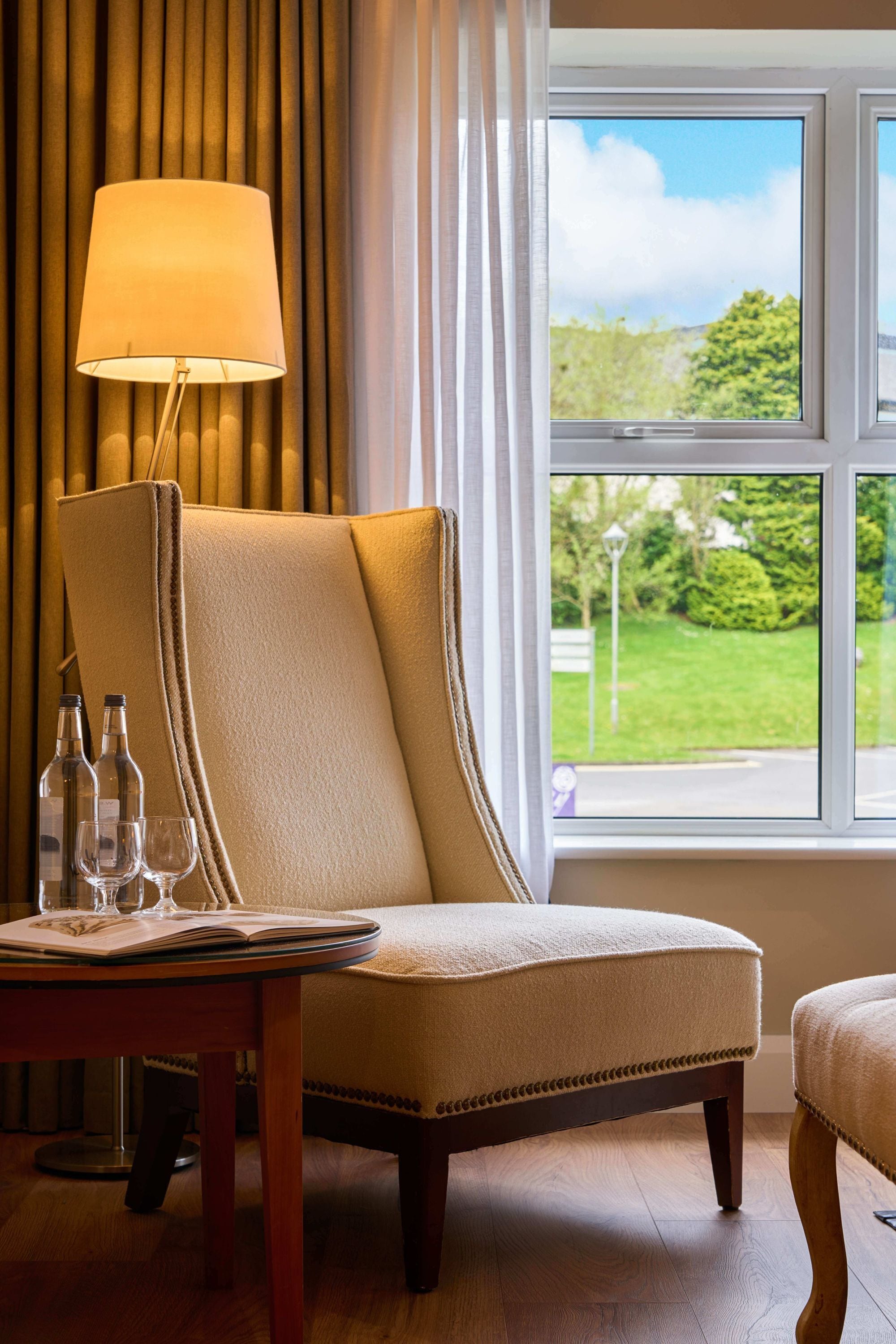 A hotel room with a large window, a chair, a table with a lamp, and a view of greenery outside.