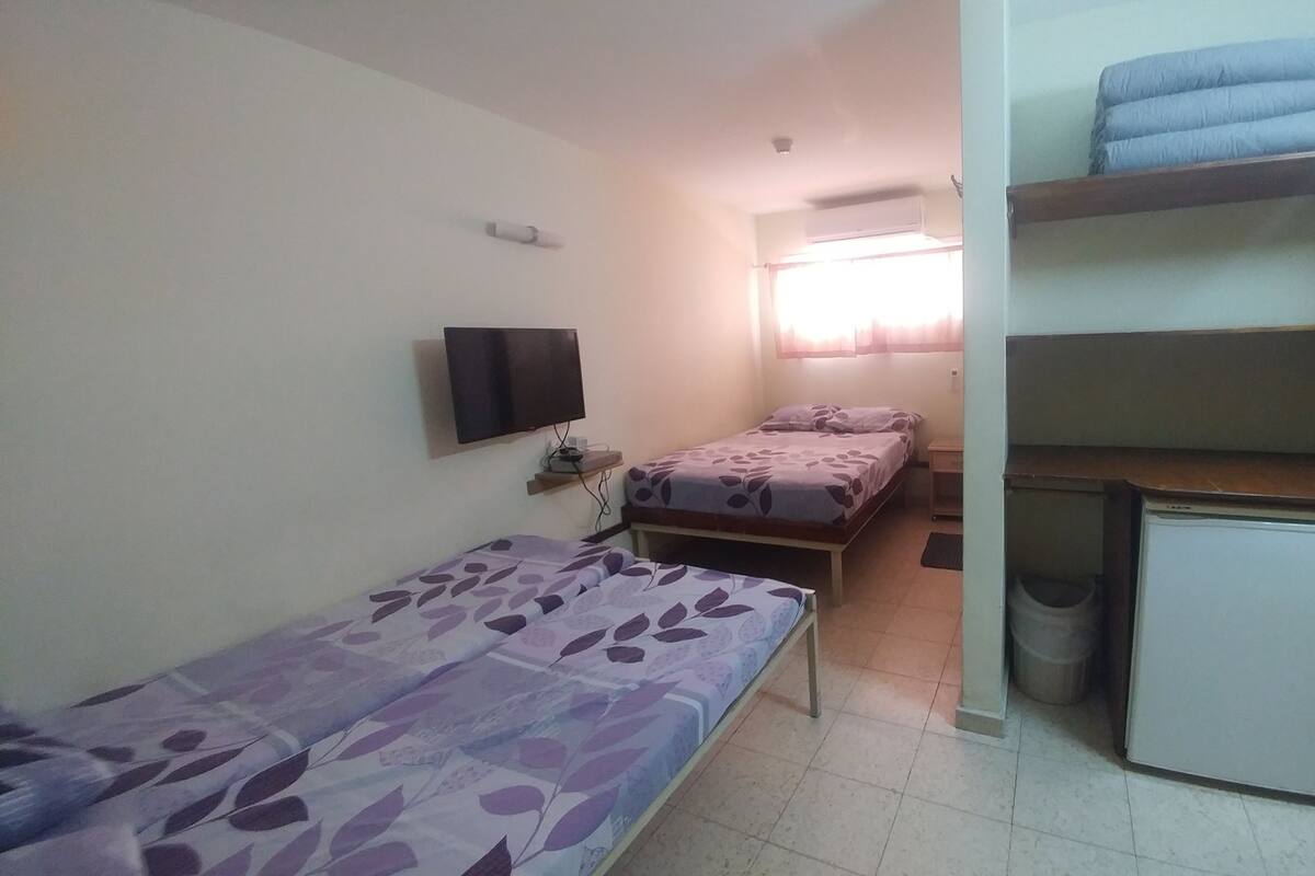 A small, simple room with two single beds, a desk, a television, and a small refrigerator.