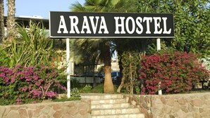 A sign for Arava Hostel with a stone wall and steps leading up to it, surrounded by lush greenery and pink flowers. - Arava Hostel (Eilat)