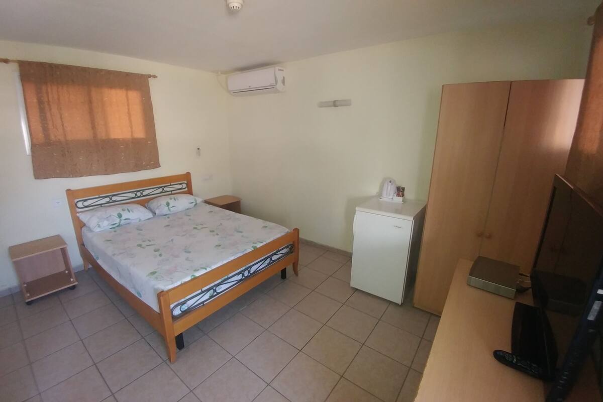 A bedroom with a bed, a small refrigerator, a wooden wardrobe, a television, and a window with a curtain.