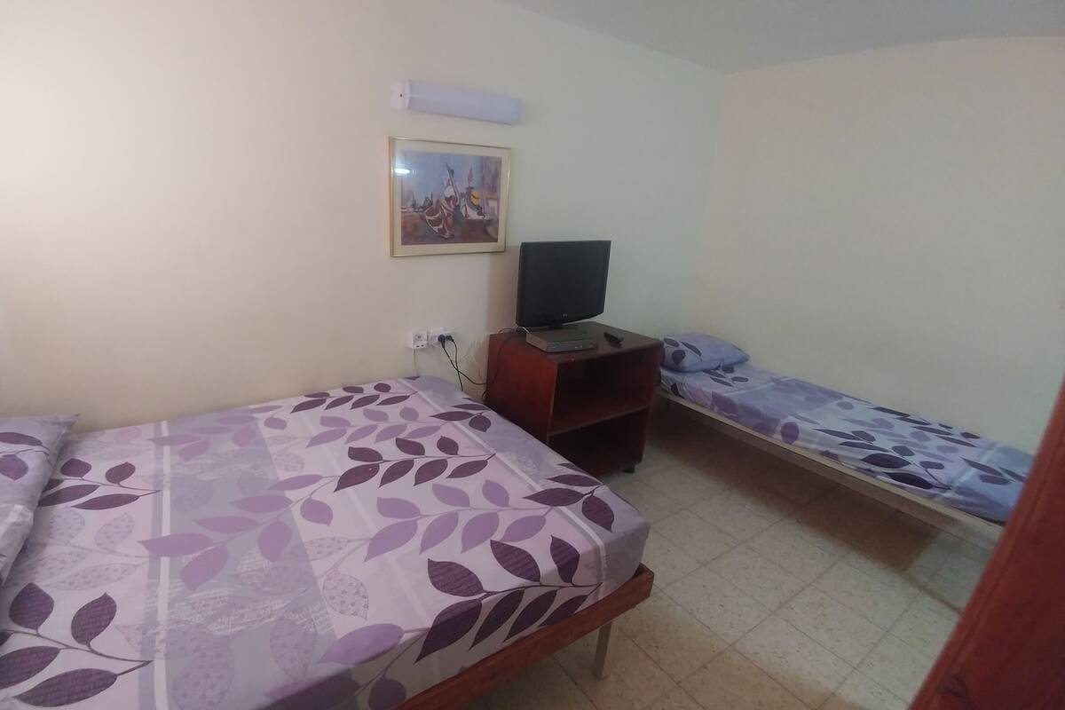 A small, simple room with two single beds, a television on a wooden stand, and a wall-mounted light fixture.
