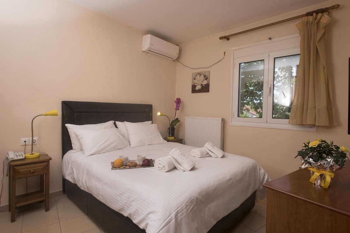 A neatly made bed with white linens, a bedside table with a lamp, a small plant, and a window with curtains.