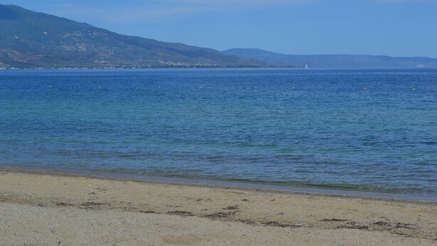 Een strand met helderblauw water, een zandstrand en op de achtergrond bergen.