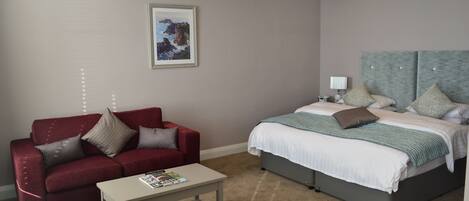 A hotel room with a red sofa, a bed with a grey headboard, a small table with a magazine, and a framed picture on the wall.