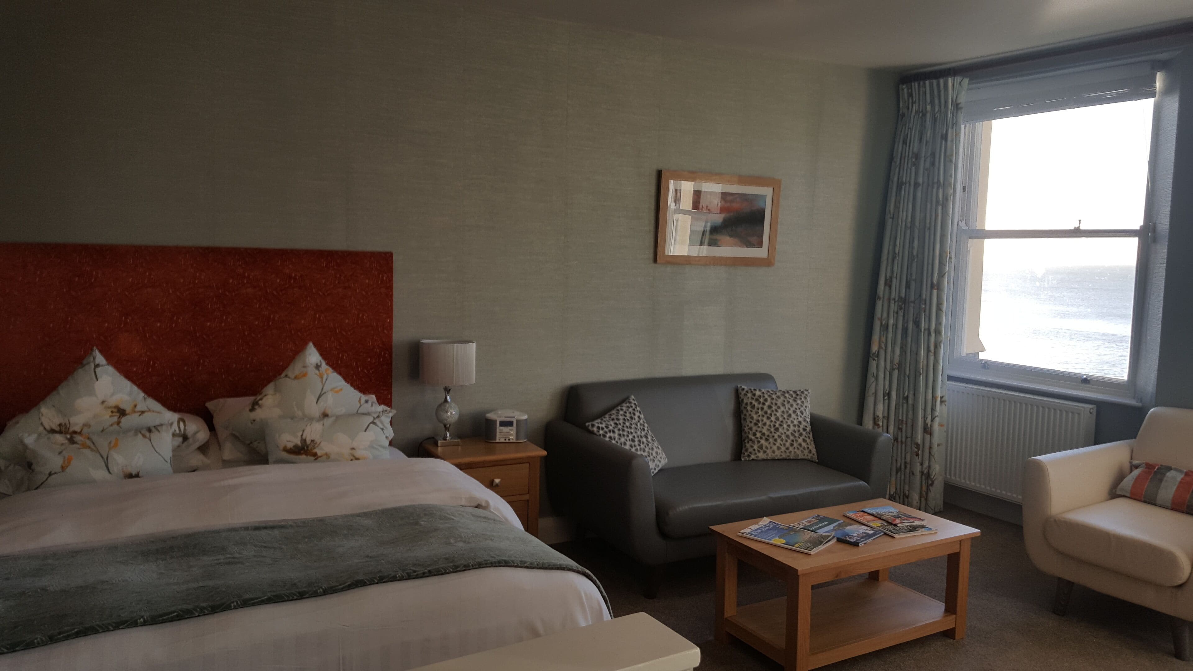Una camera d'albergo con un letto, un divano grigio, un tavolino in legno, un comodino con una lampada e una finestra con tende.