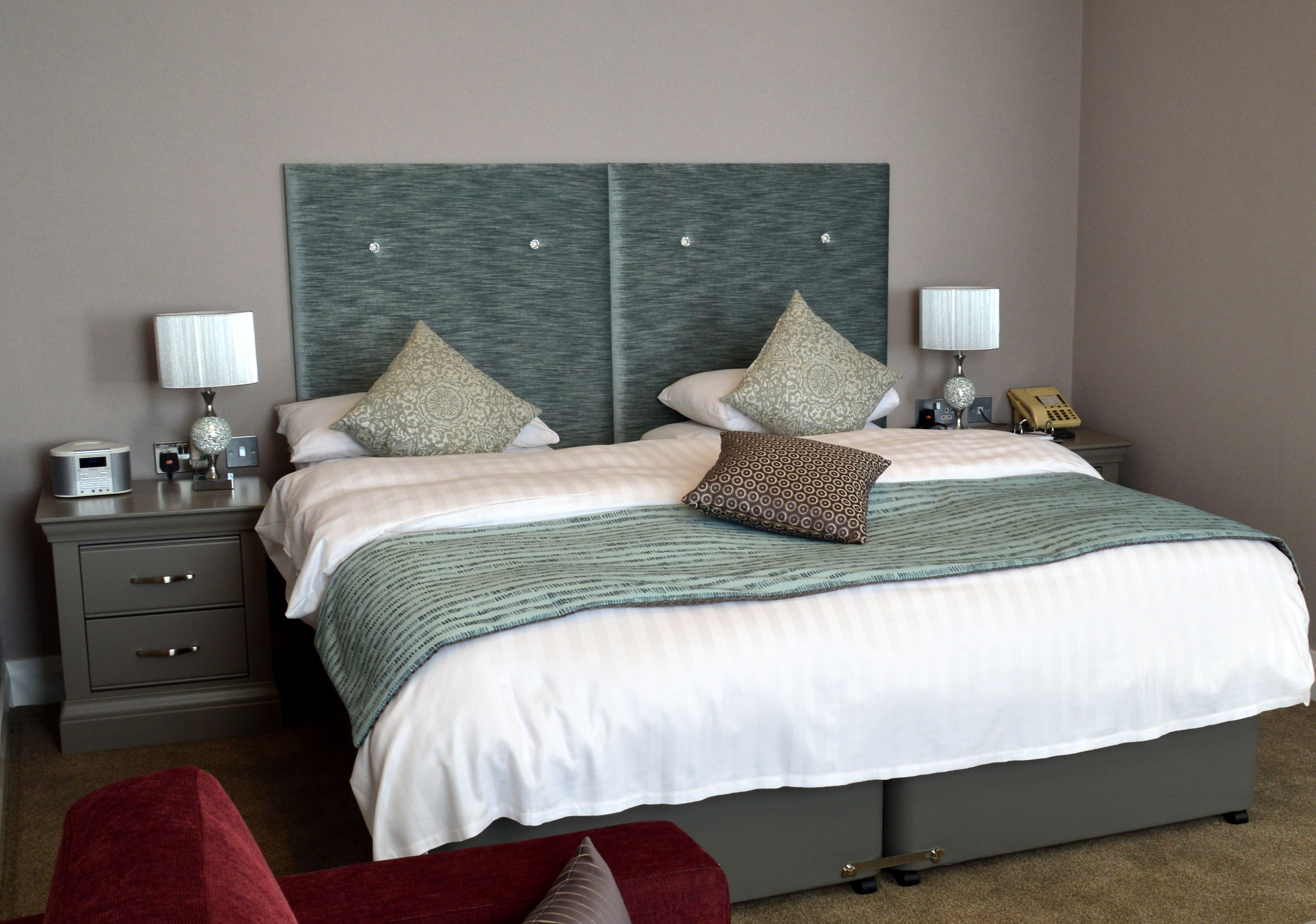 A neatly made bed with a grey headboard, two bedside tables with lamps, a telephone, and a red chair.