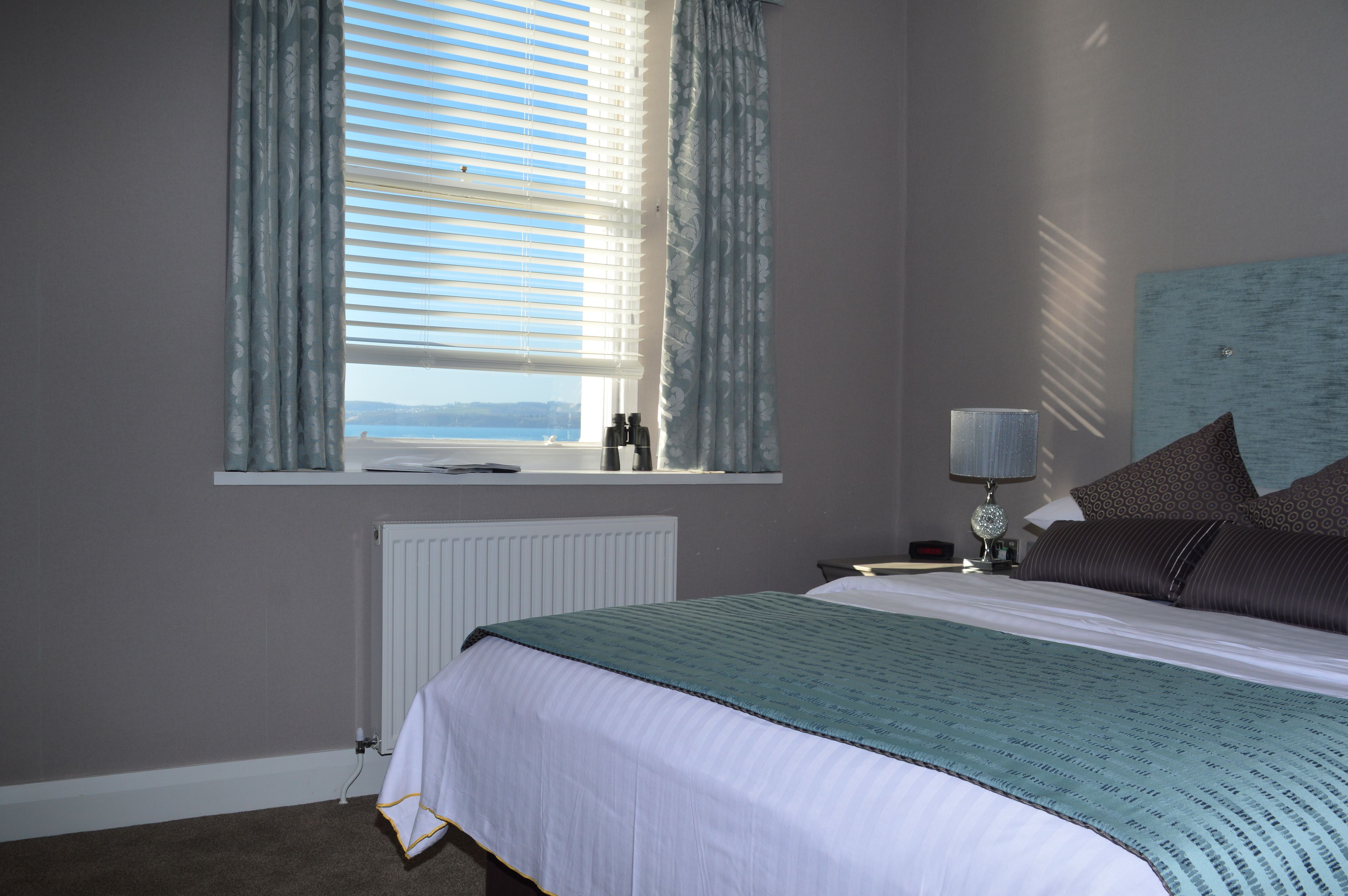 A bedroom with a large bed, a window with curtains, a bedside table with a lamp, and a view of the sea.