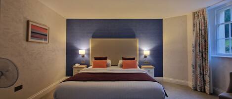 A hotel room with a large bed, two bedside tables with lamps, a wall-mounted fan, a window with curtains, and a painting on the wall.