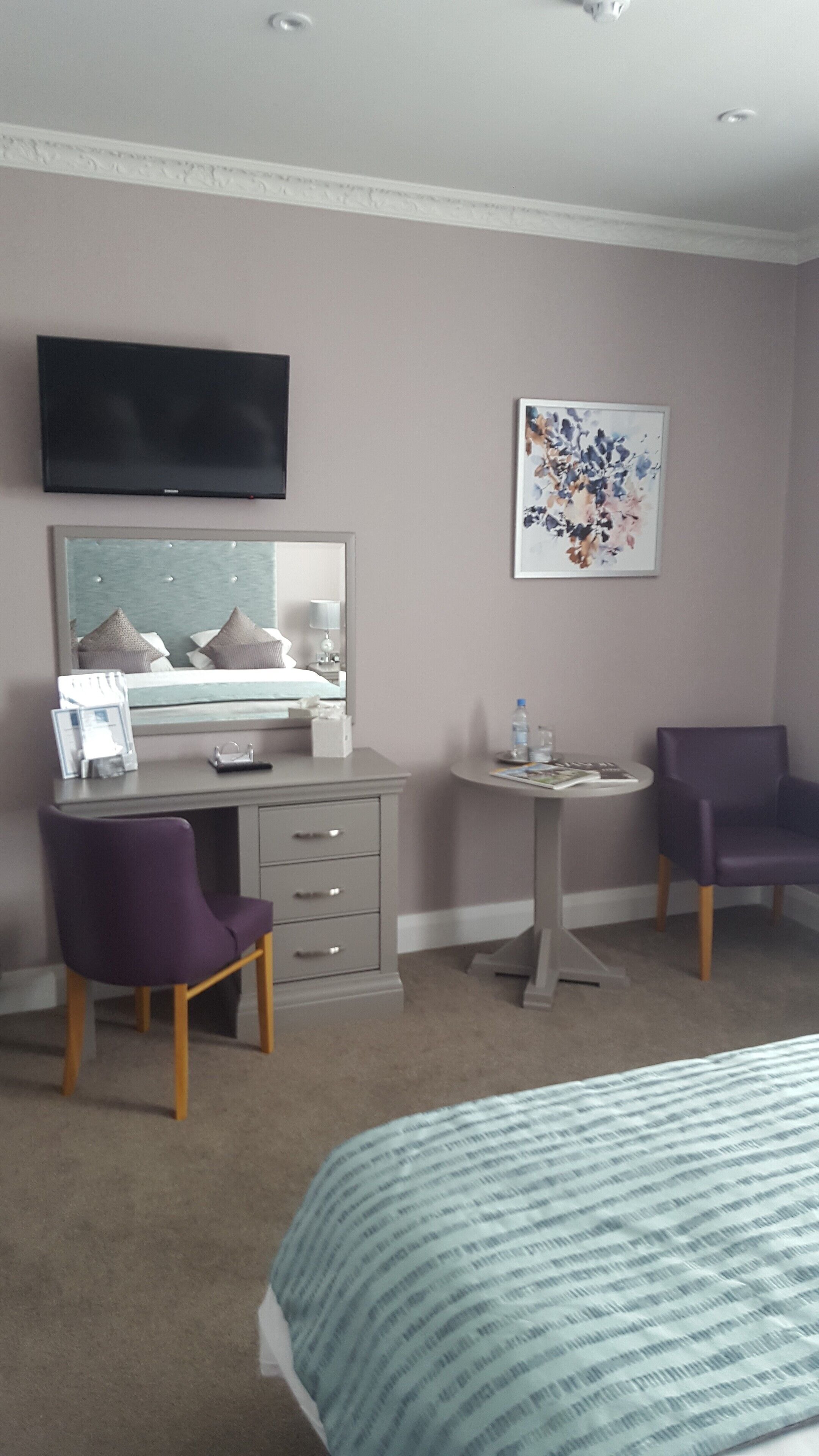 A hotel room with a bed, a desk with a chair, a television, a small table with a chair, and a framed picture on the wall.