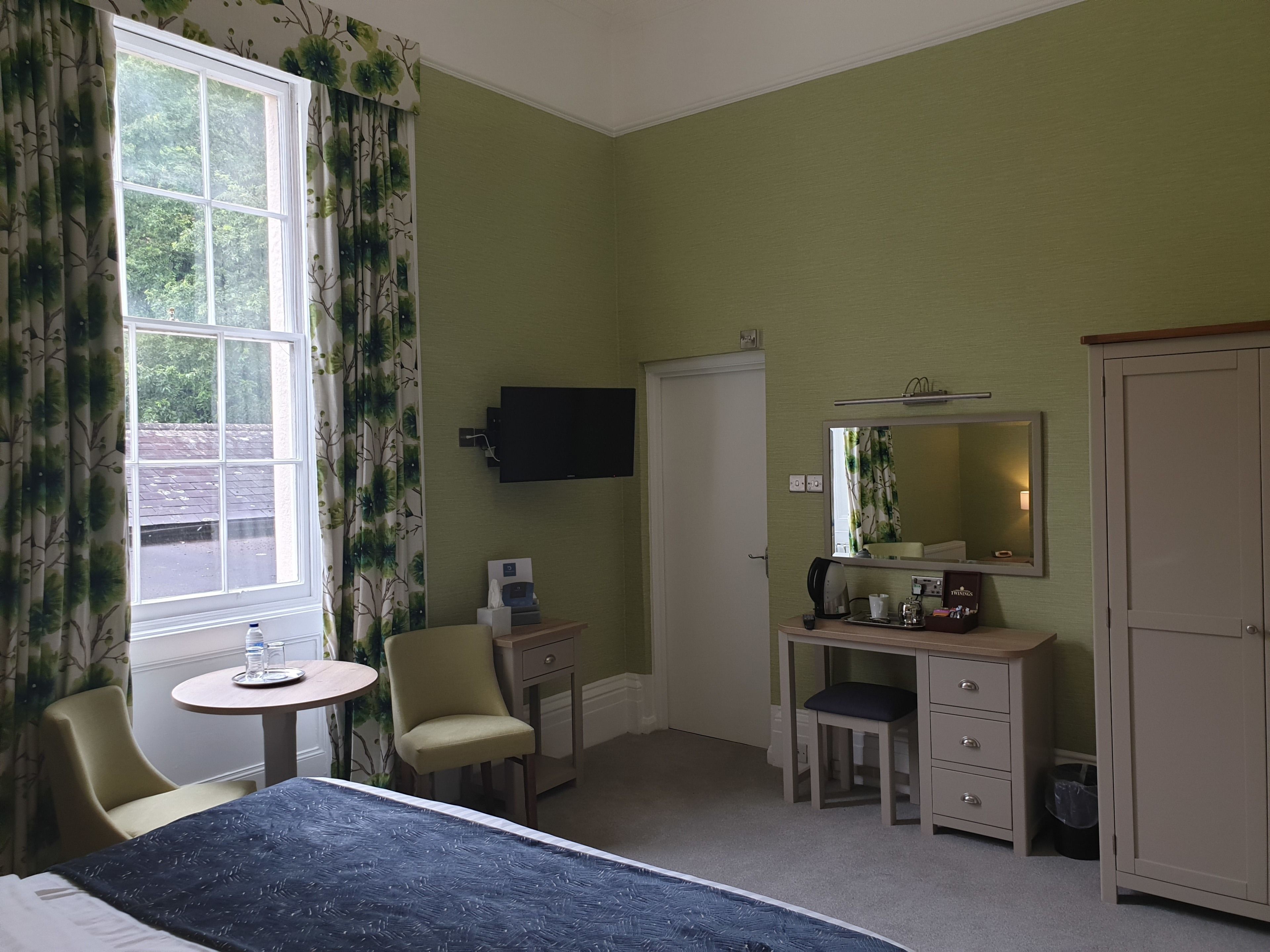 A hotel room with a bed, a desk, a chair, a small table, a TV, a mirror, and a wardrobe.