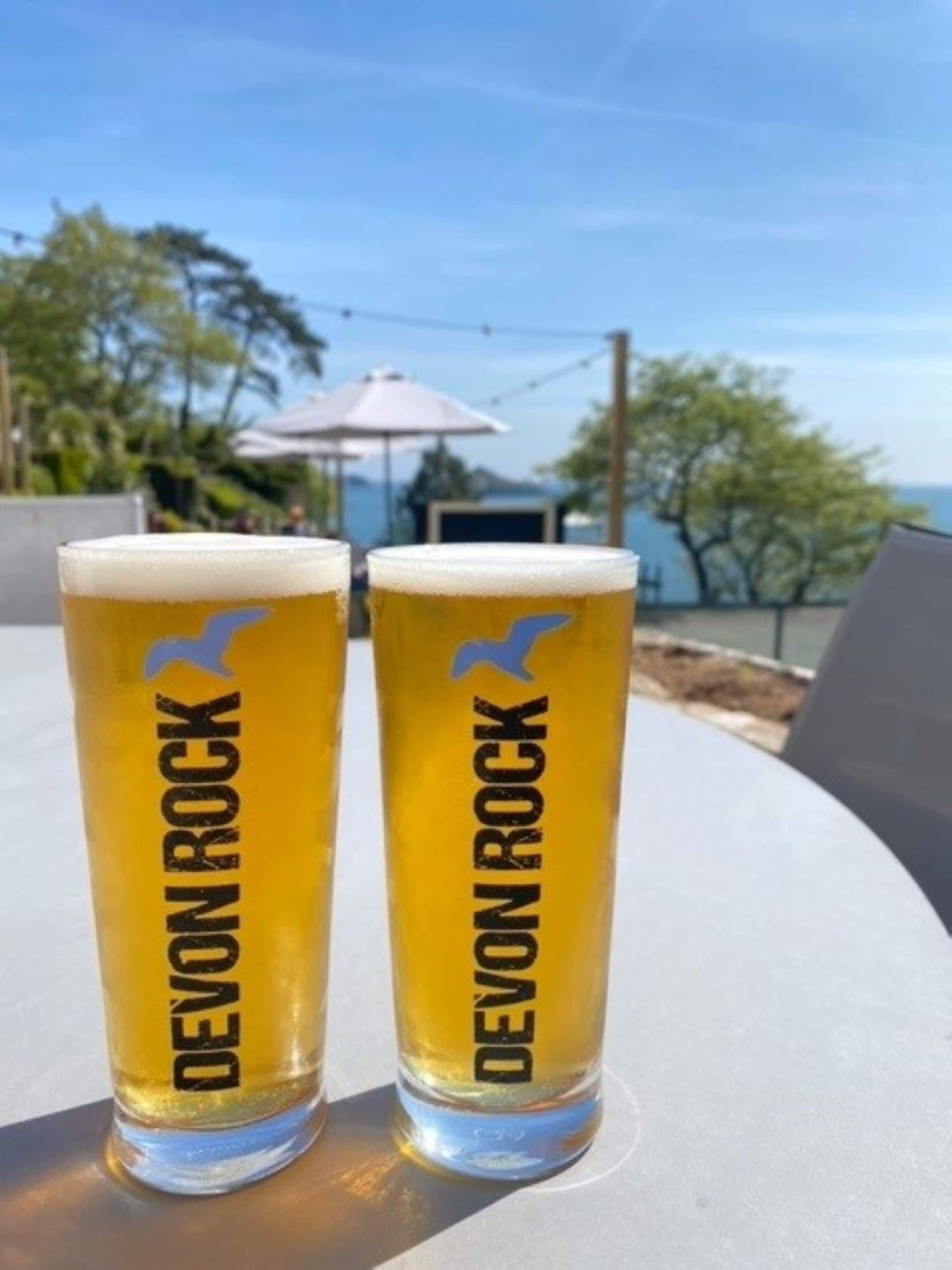 Zwei Biergläser mit 'Devon Rock'-Branding auf einem Tisch im Freien.