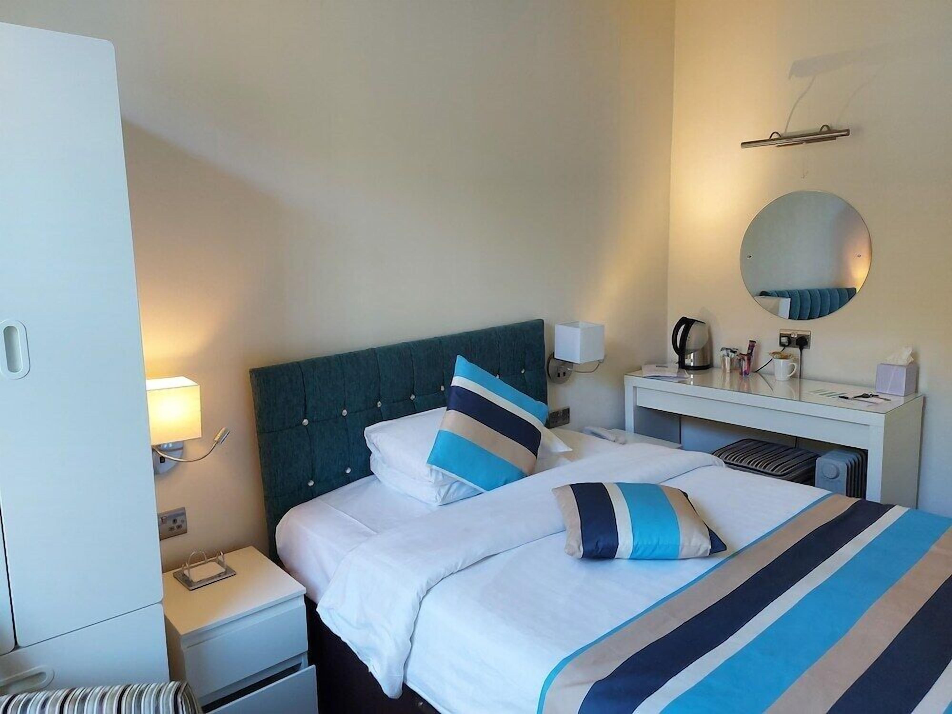 A hotel room with a bed, bedside table, lamp, mirror, and a small desk with a coffee maker.
