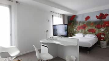 A modern hotel room with a white desk, a bed, a TV, and a wall mural of red flowers.