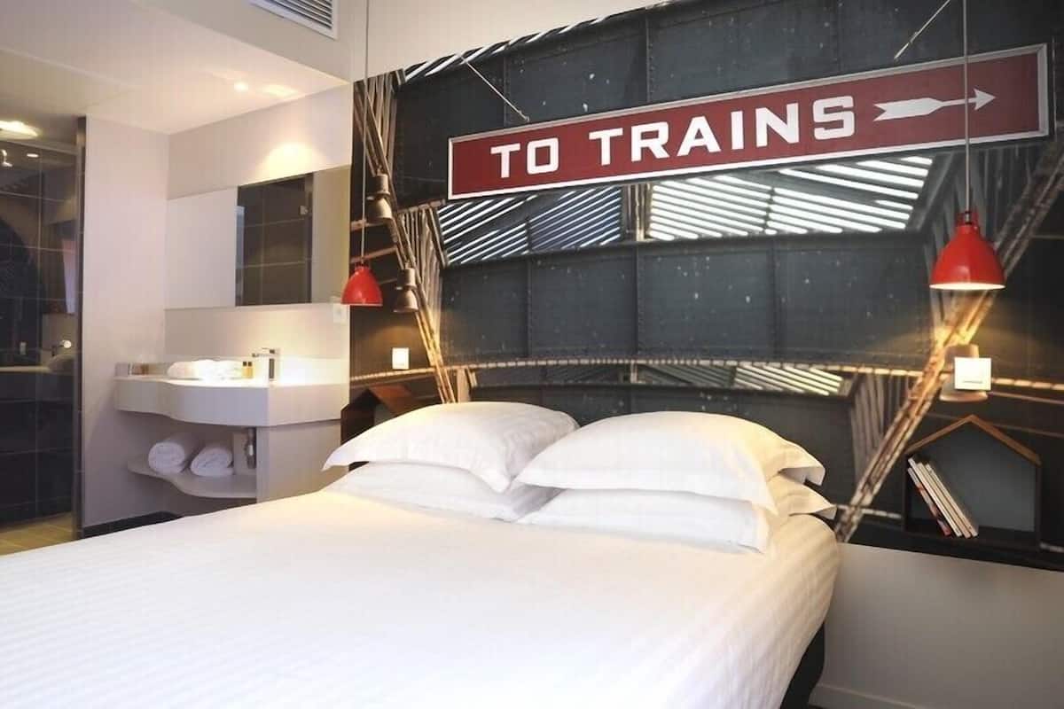 A modern hotel room with a bed, a nightstand, a mirror, and a sign that reads "TO TRAINS".
