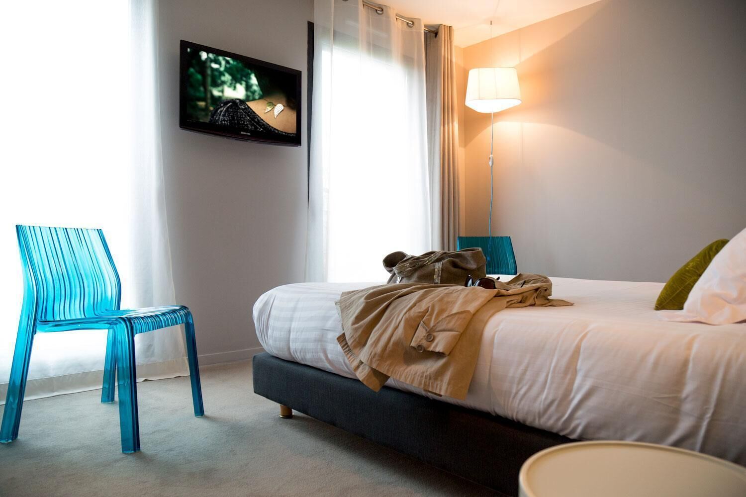 A hotel room with a bed, a TV mounted on the wall, a chair, and a bedside lamp.