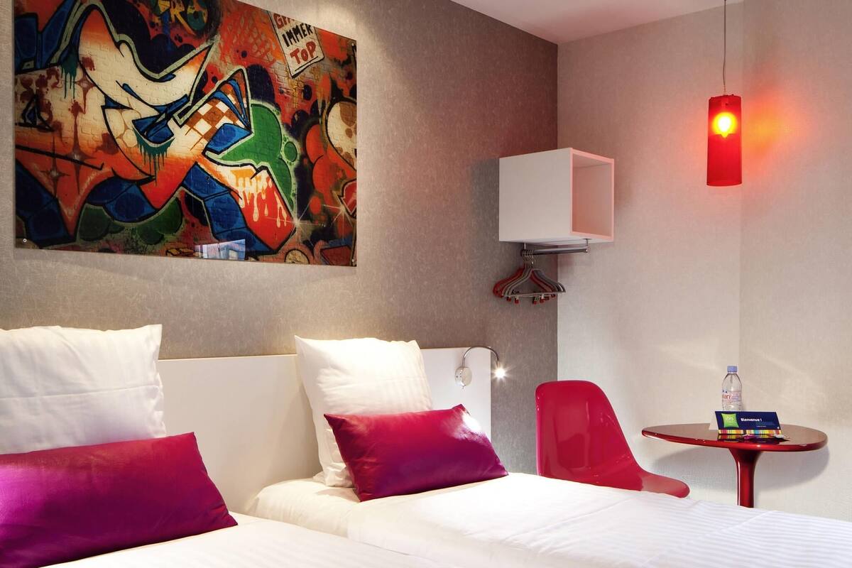 A hotel room with a bed, pillows, a red chair, a small table with a bottle, and a wall with graffiti art.
