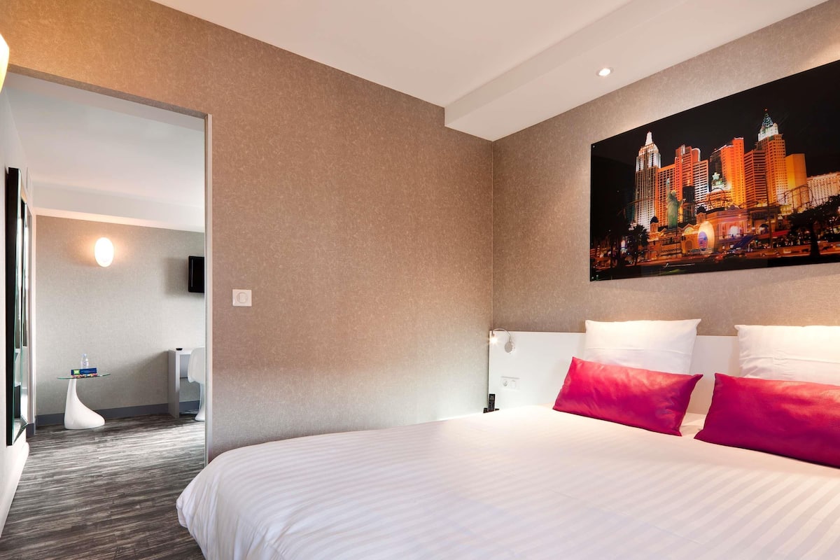 Une chambre d’hôtel avec un grand lit, un tableau représentant un paysage urbain accroché au mur et une vue sur la ville de nuit.