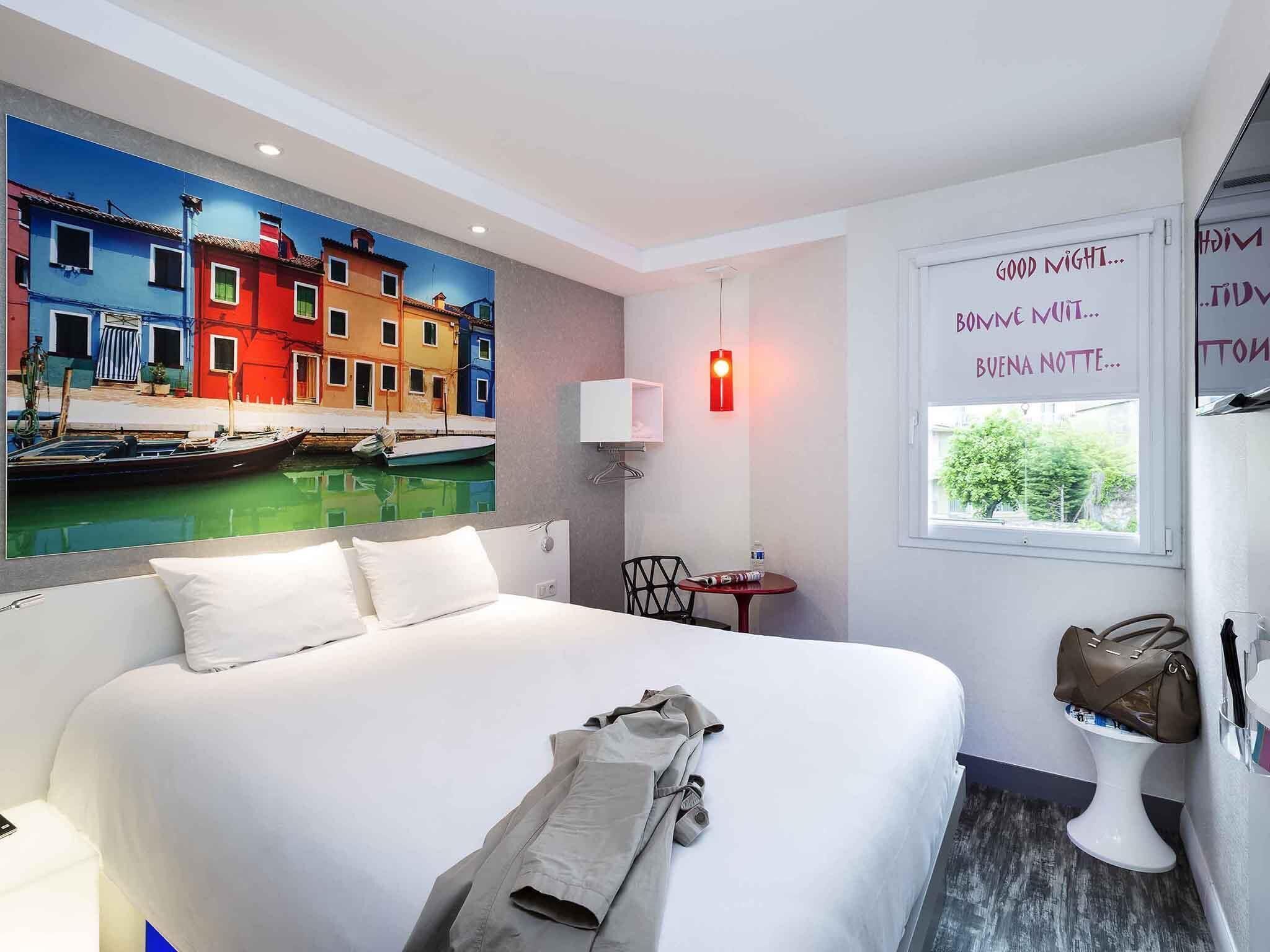 Ein Hotelzimmer mit Bett, einem Gemälde bunter Gebäude, einem kleinen Tisch mit Stuhl, einer Nachttischlampe und einem Fenster mit Blick.
