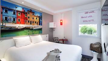 A hotel room with a bed, a painting of colorful buildings, a small table with a chair, a bedside lamp, and a window with a view.