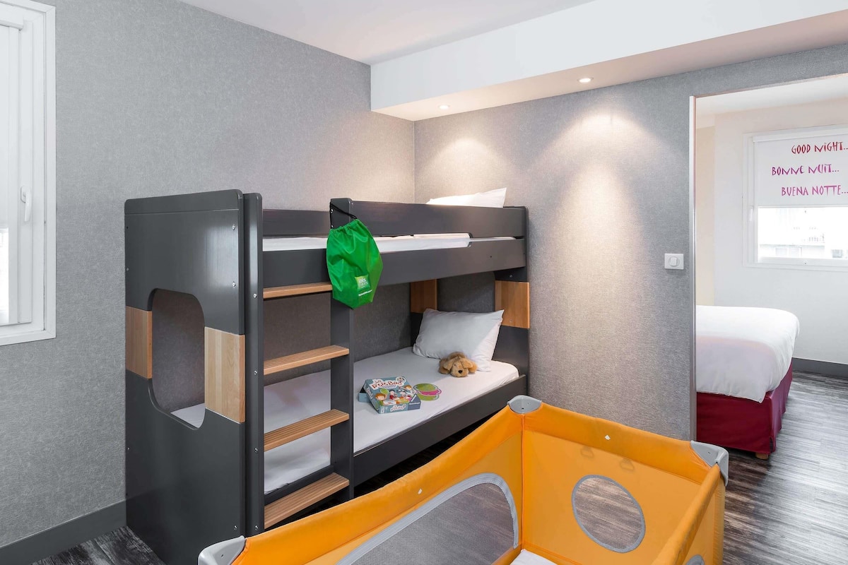 Un lit superposé avec un sac vert accroché sur le côté, un petit espace de jeux avec un parc pour enfants jaune, et une fenêtre ornée d’une pancarte rédigée en plusieurs langues.