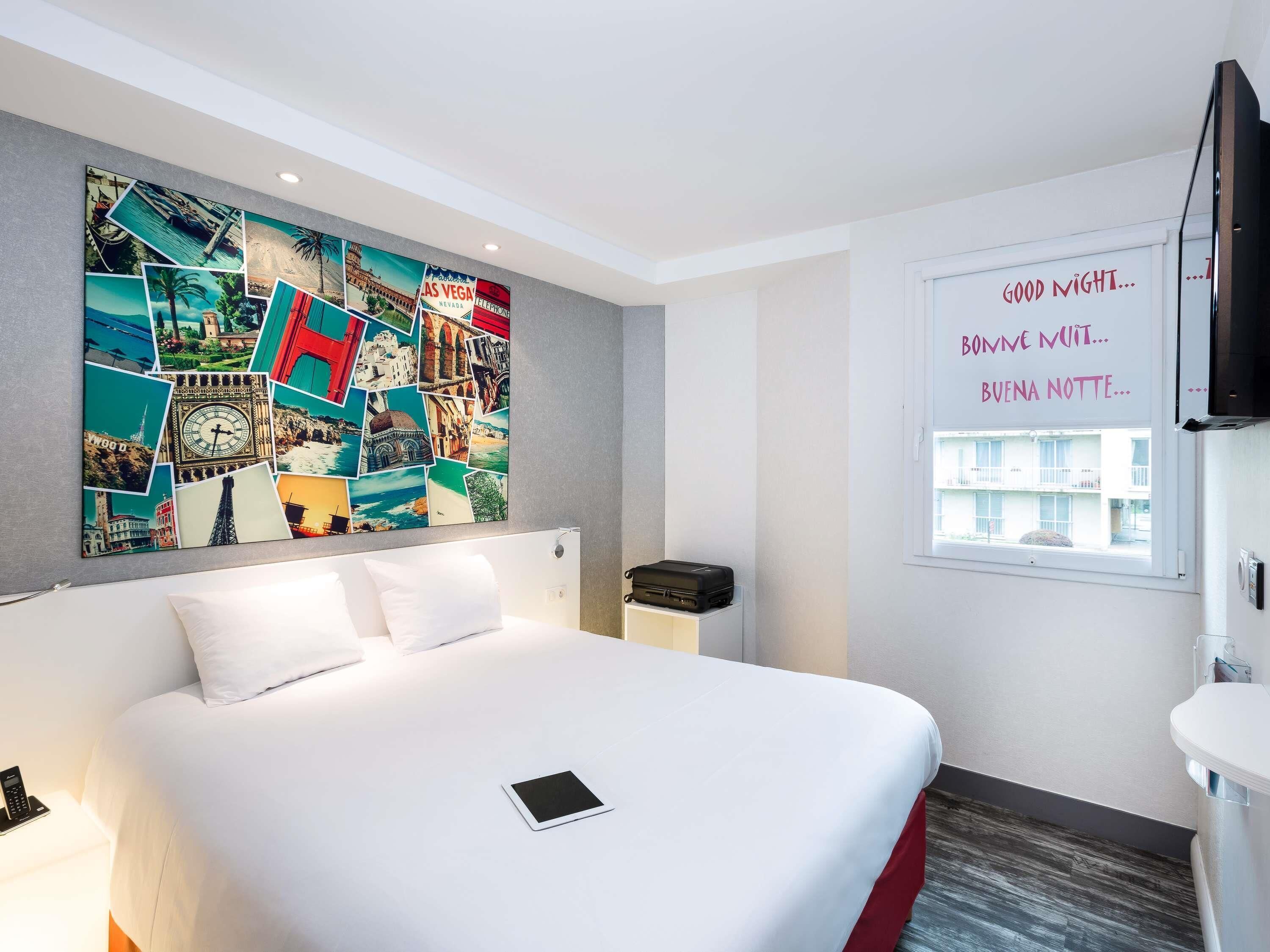 Quarto de hotel com uma grande colagem na parede, uma cama, uma mesinha de cabeceira, uma televisão e uma janela com vista para edifícios.