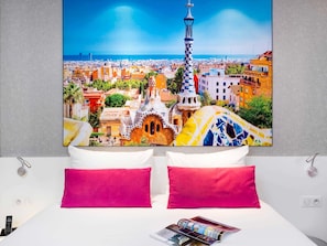 A hotel room with a bed, two pillows, a magazine, and a large cityscape painting on the wall.