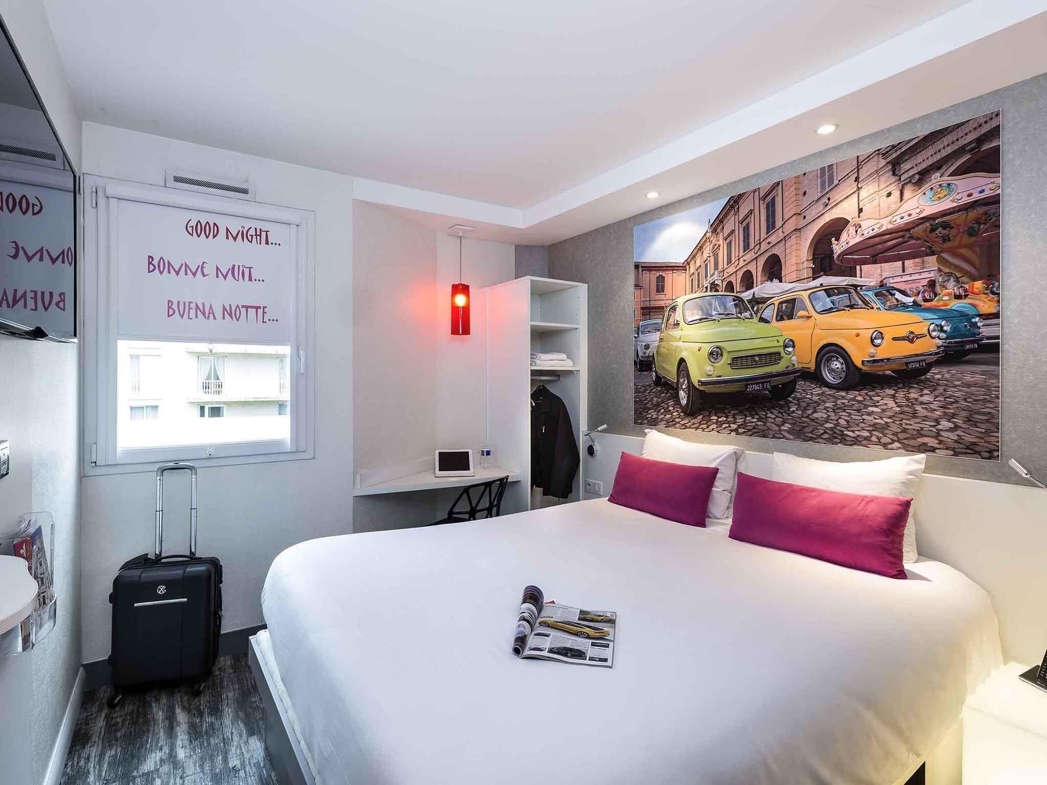 A hotel room with a bed, a suitcase, a desk, a chair, a window with a view of a street, and a wall with a painting of vintage cars.