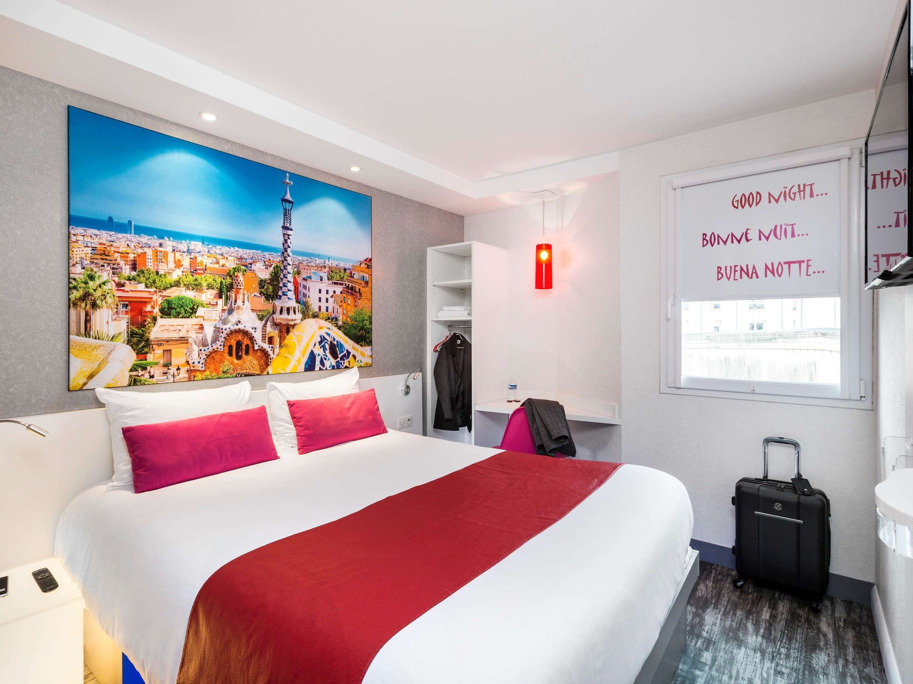 A hotel room with a large bed, a cityscape painting, a wardrobe, a suitcase, and a window with a red light.