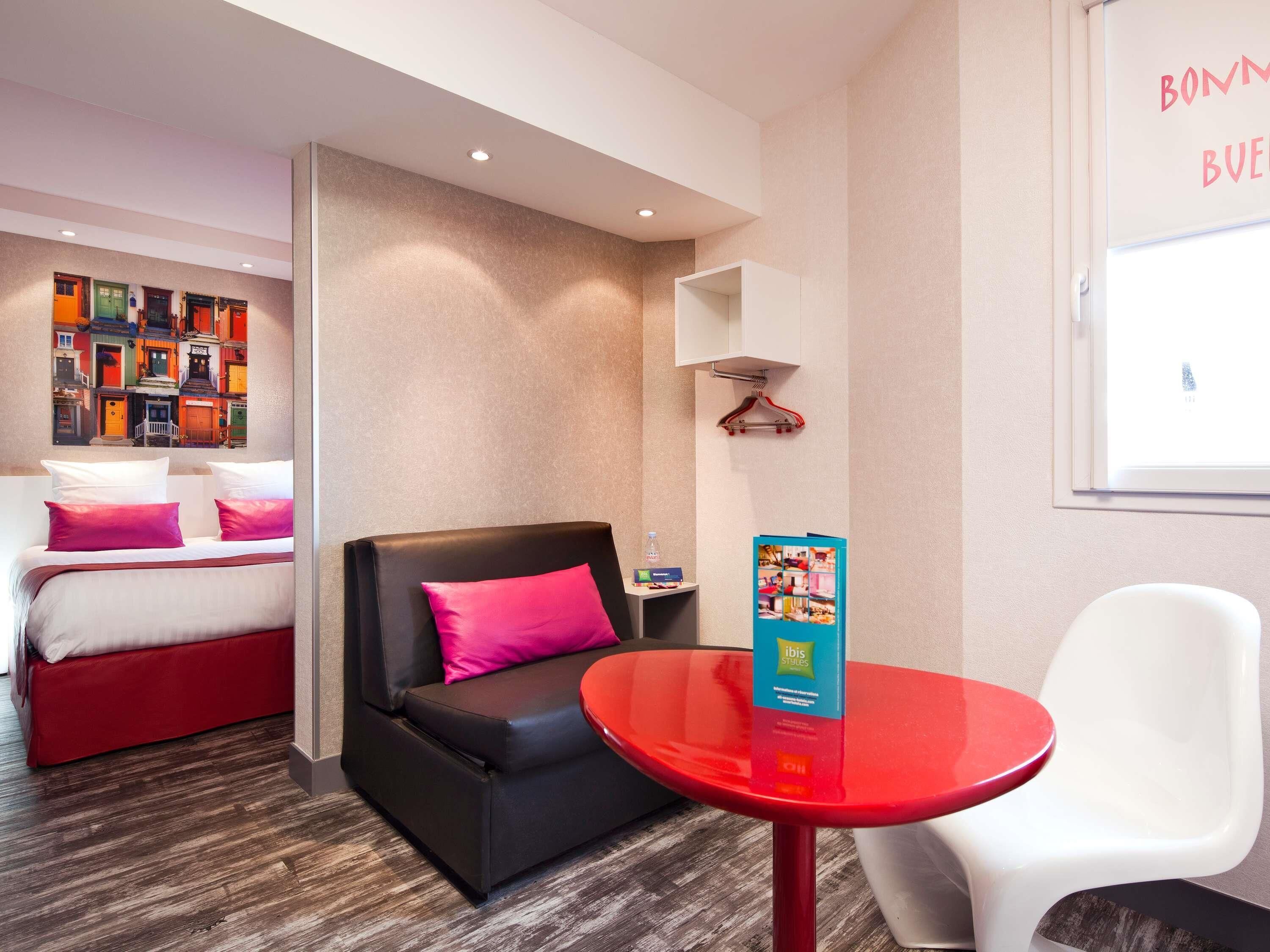 A hotel room with a bed, a black sofa, a red table, and a white chair.