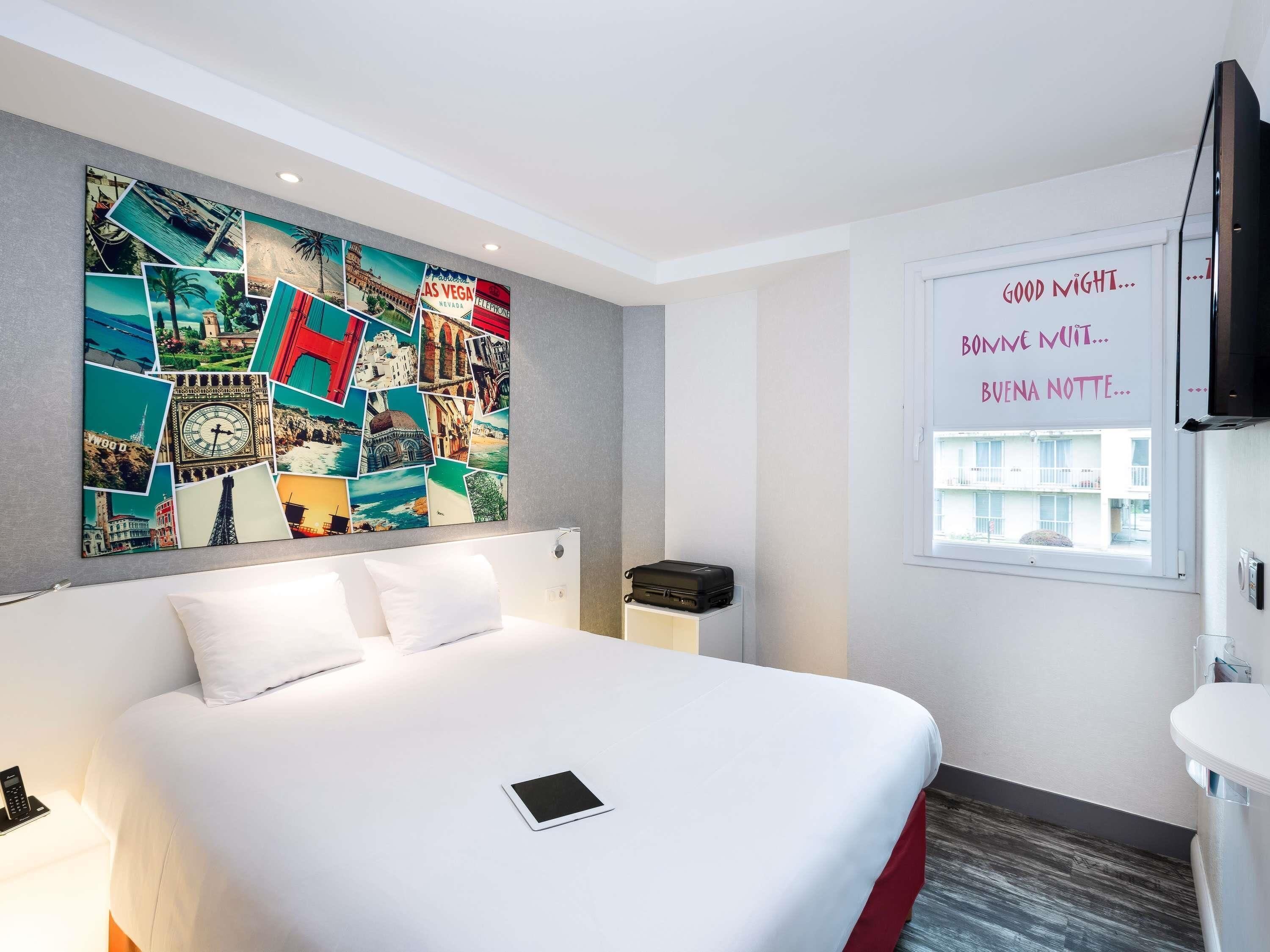 Une chambre d’hôtel avec un lit, un collage artistique au mur, une fenêtre donnant sur un paysage agréable et une table de chevet avec une télécommande.