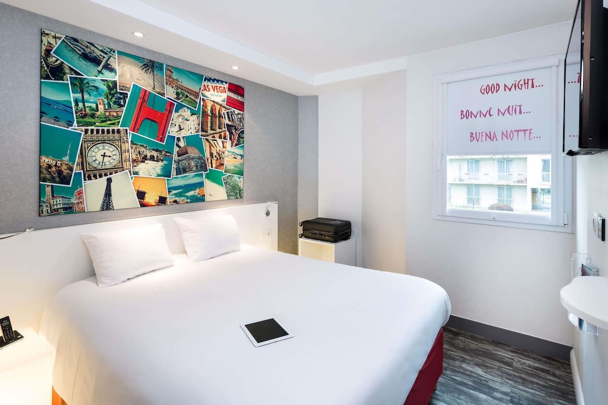 Une chambre d’hôtel avec un lit, un collage artistique au mur, une fenêtre donnant sur un paysage agréable et une table de chevet avec une télécommande.