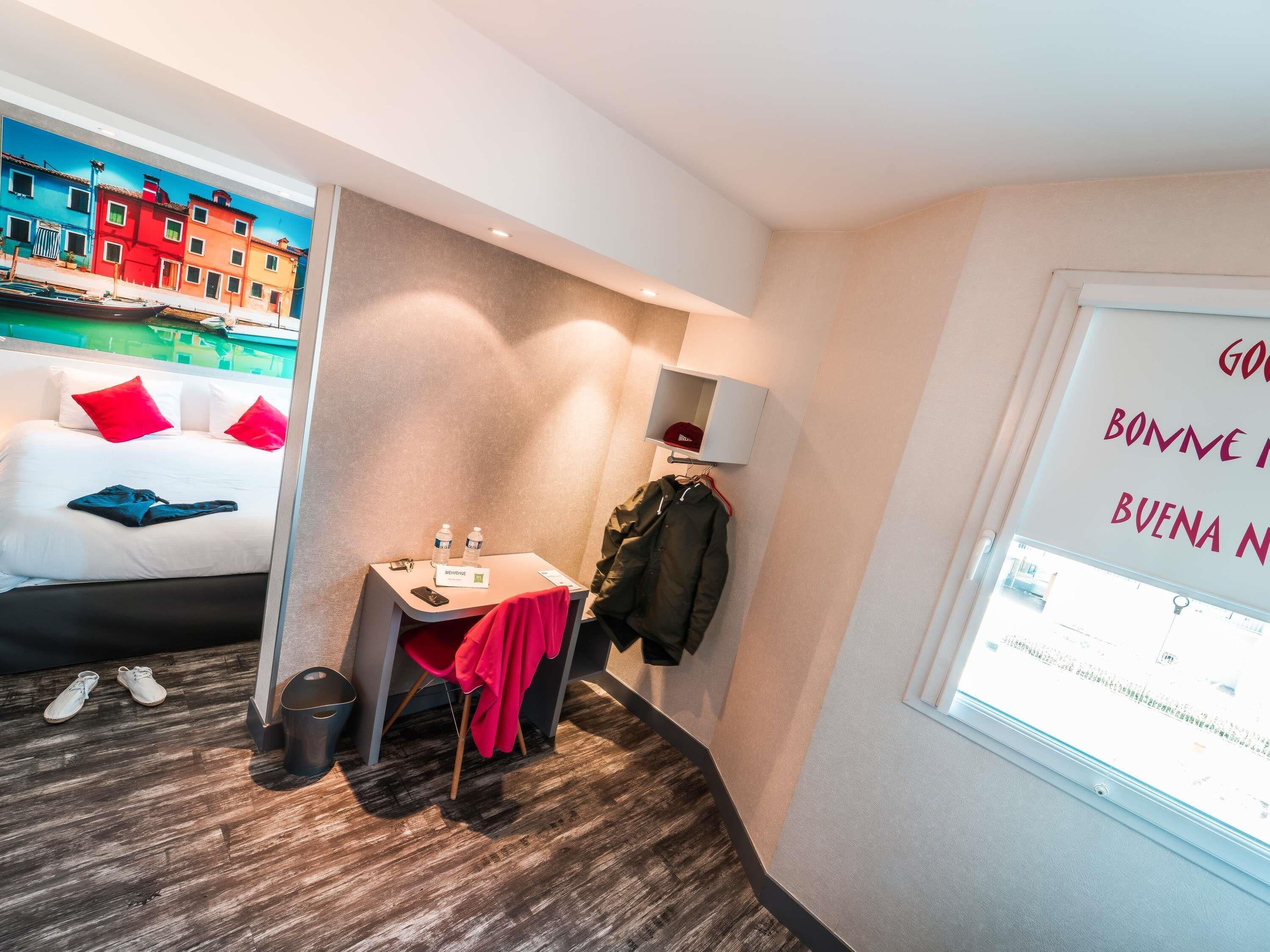 Habitación de hotel con cama, escritorio, silla, perchero y una pared que ofrece una vista parcial de una calle colorida.