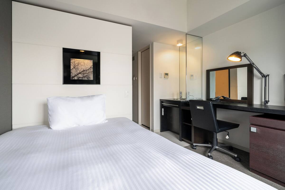 A hotel room with a bed, a desk, a chair, a mirror, and a picture on the wall.