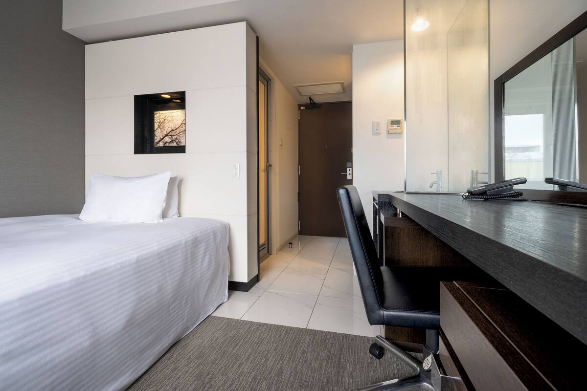 A hotel room with a bed, a desk with a chair, a large mirror, and a door leading to another room.