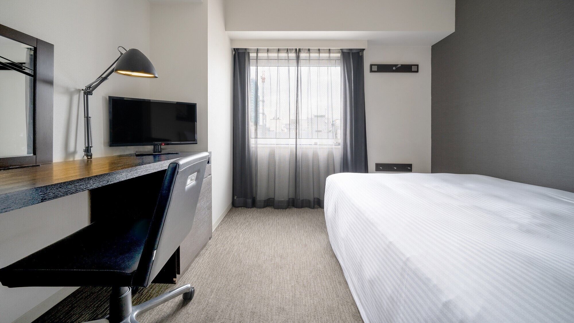 A hotel room with a bed, a desk with a computer monitor, a chair, a desk lamp, and a window with sheer curtains.