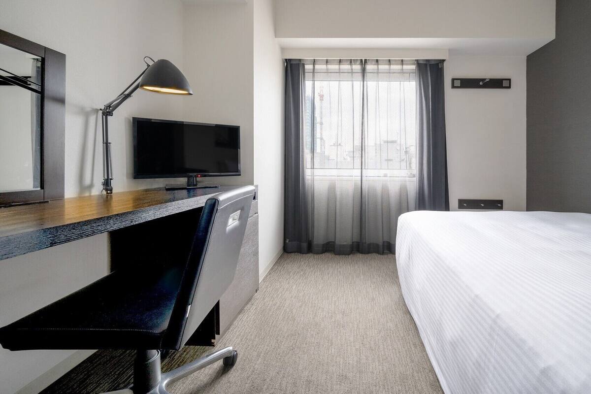 A hotel room with a bed, a desk with a computer monitor, a chair, a desk lamp, and a window with sheer curtains.