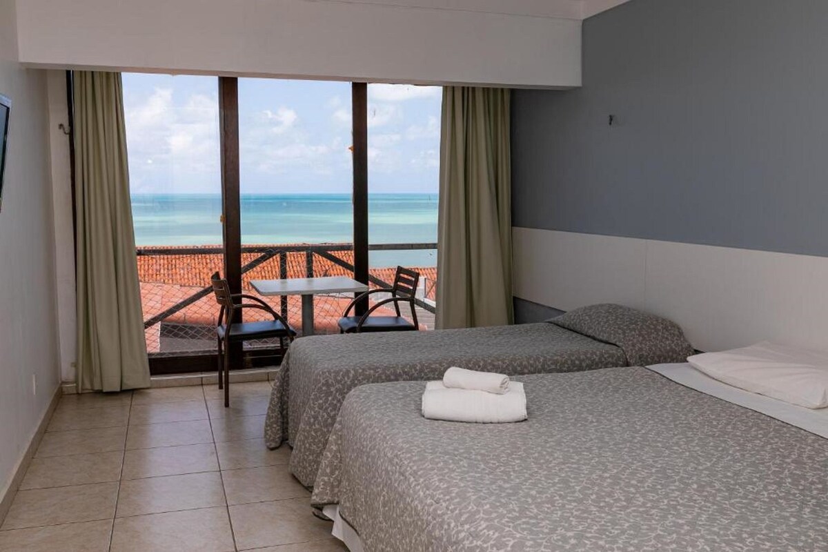 Quarto de hotel com duas camas, uma varanda com mesa e cadeiras, e vista para o mar.