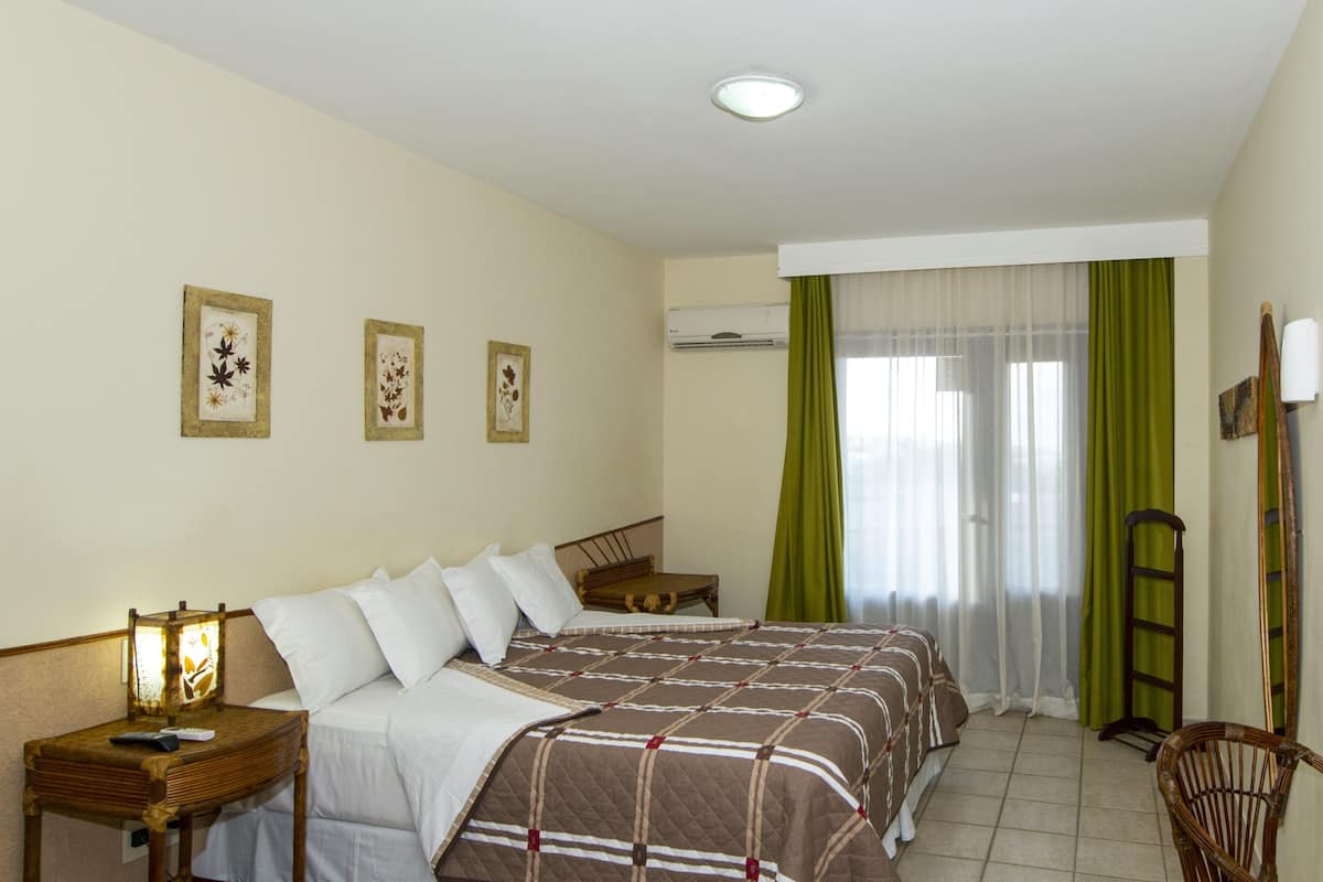 A hotel room with a large bed, a bedside table with a lamp, a chair, a mirror, and a window with curtains.
