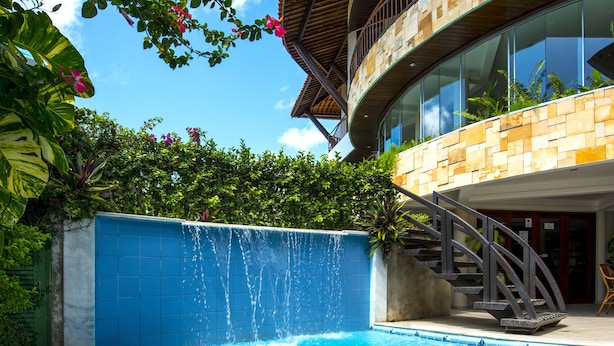 A modern pool area with a clear blue pool, a thatched roof structure, and a stone building with a balcony. - Divi Divi Praia Hotel (Natal)