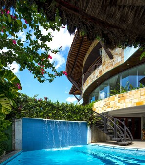 A modern pool area with a clear blue pool, a thatched roof structure, and a stone building with a balcony.