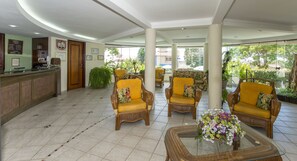 A lobby with a reception desk, wicker chairs, a glass coffee table, and a view of greenery outside.