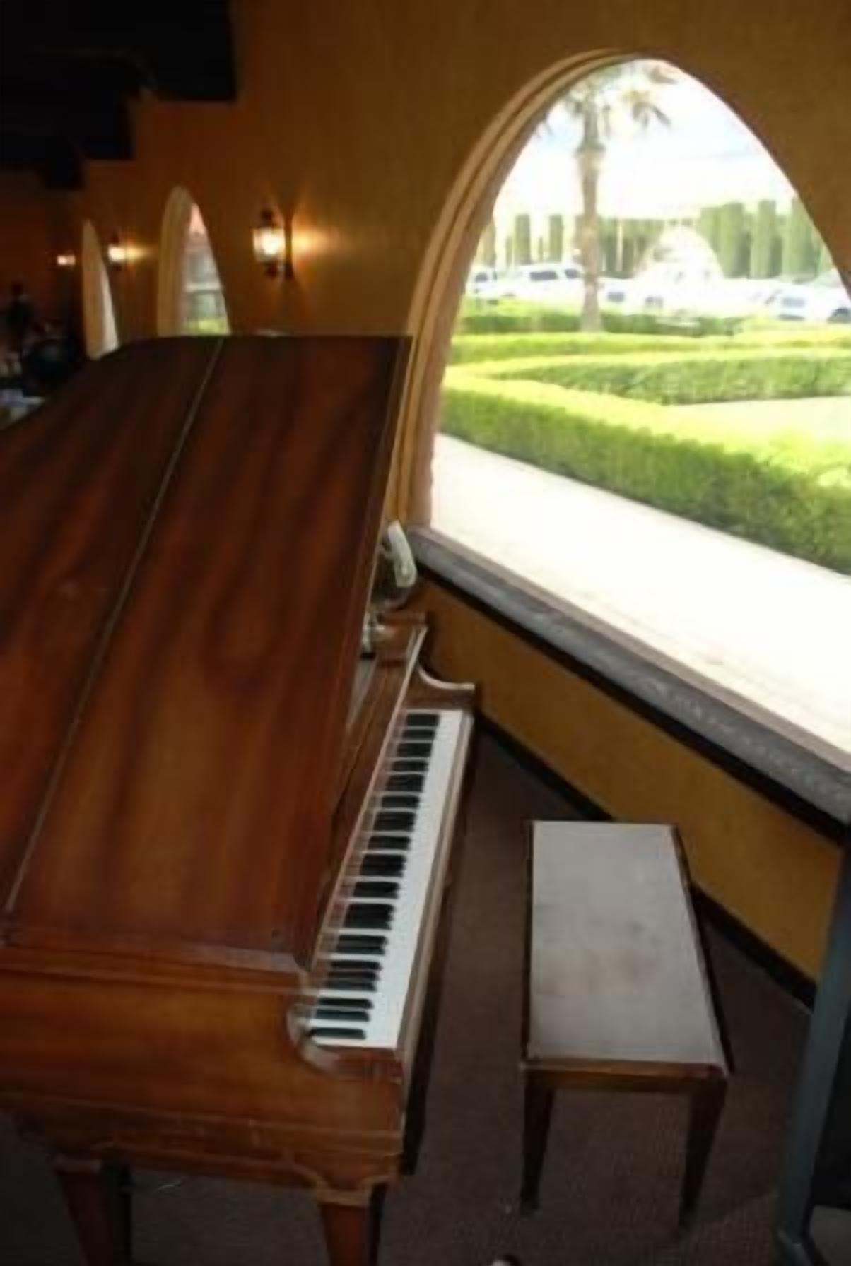 Un piano à queue se trouve dans une pièce dotée d’une grande fenêtre en arc qui donne sur une pelouse bien entretenue et des voitures garées.