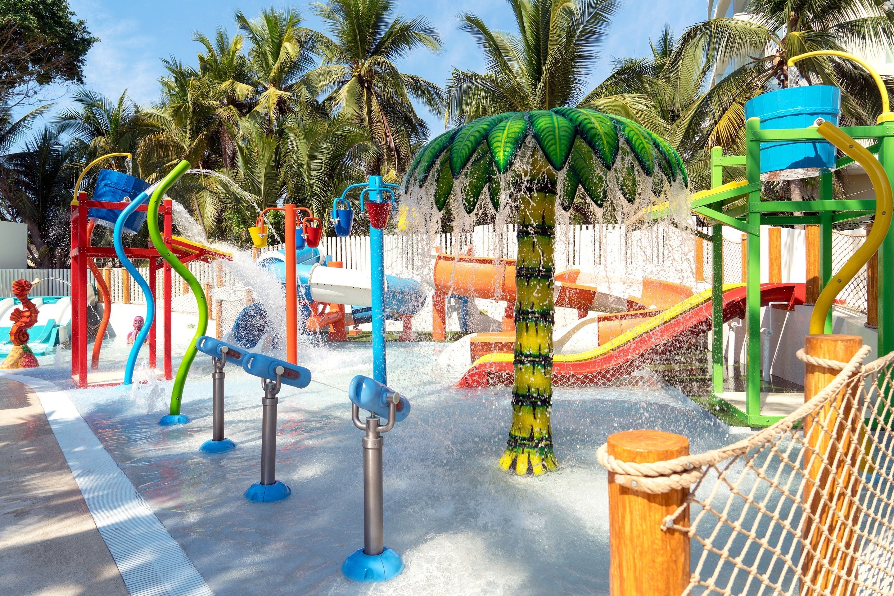 A water play area with slides and water jets, surrounded by palm trees.