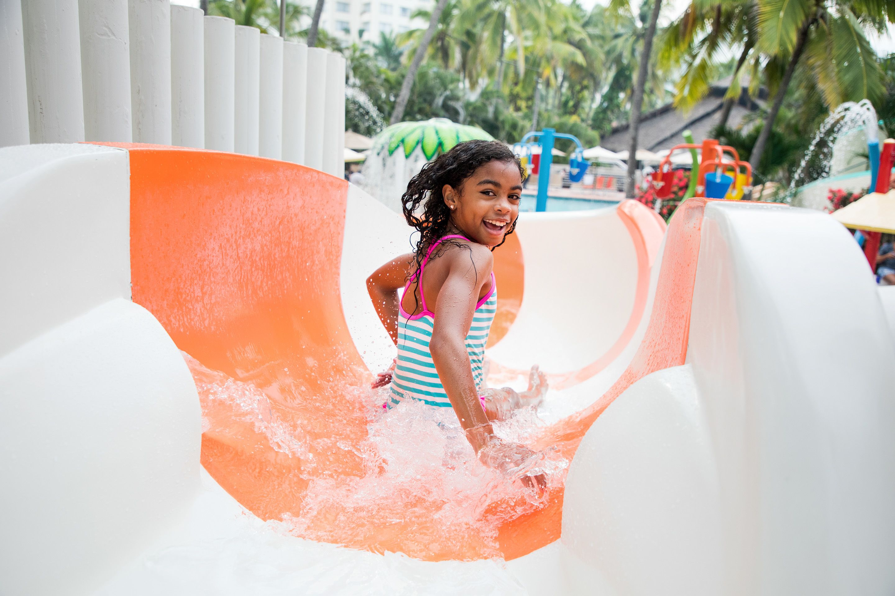 Un niño resbalando por un tobogán acuático con diseño blanco y naranja, rodeado de palmeras y una zona de piscina.